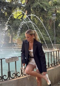 Veste noire à rayures sur une robe blanche, ceinture noire et chaussures blanches. Tenant un petit sac, debout à côté d'une fontaine entourée de verdure.