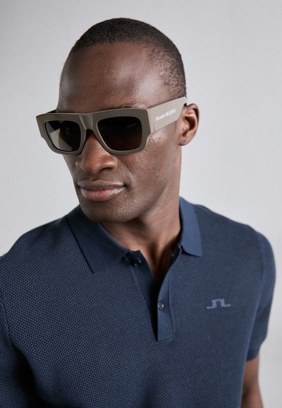Gafas de sol de plástico marrón con lentes cuadrados y montura gruesa, con la marca "Alexander McQueen" en el lateral, llevadas con un polo azul marino.