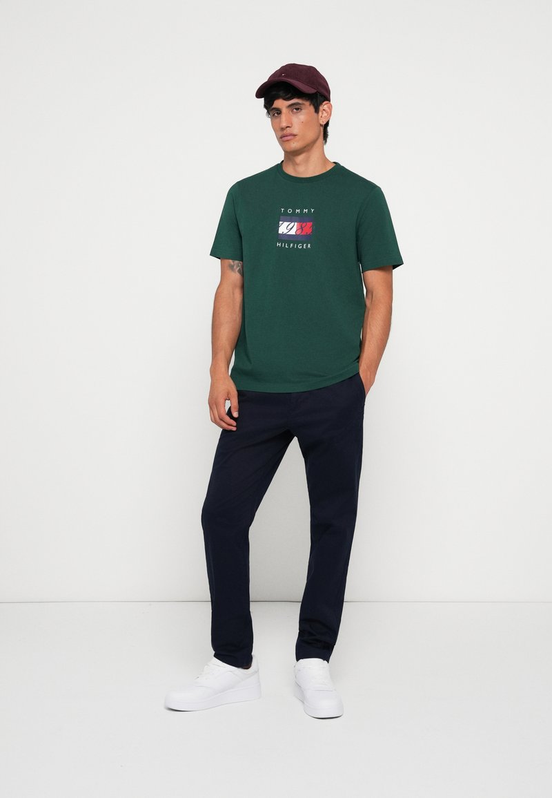 Una camiseta de algodón verde presenta un logo impreso en rojo, blanco y azul, combinada con pantalones marinos y zapatillas blancas, llevada con una gorra color burdeos.