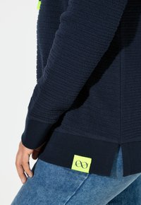 Sudadera azul marino texturizada con puños y dobladillo acanalados. Cuenta con una etiqueta amarilla brillante con un símbolo de infinito. Combinada con vaqueros azules.