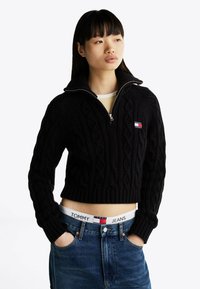 Maglione corto nero a trecce con colletto a mezzo zip, caratterizzato da una piccola toppa con logo. Abbinato a jeans a vita alta con marchio visibile sul cinturino.