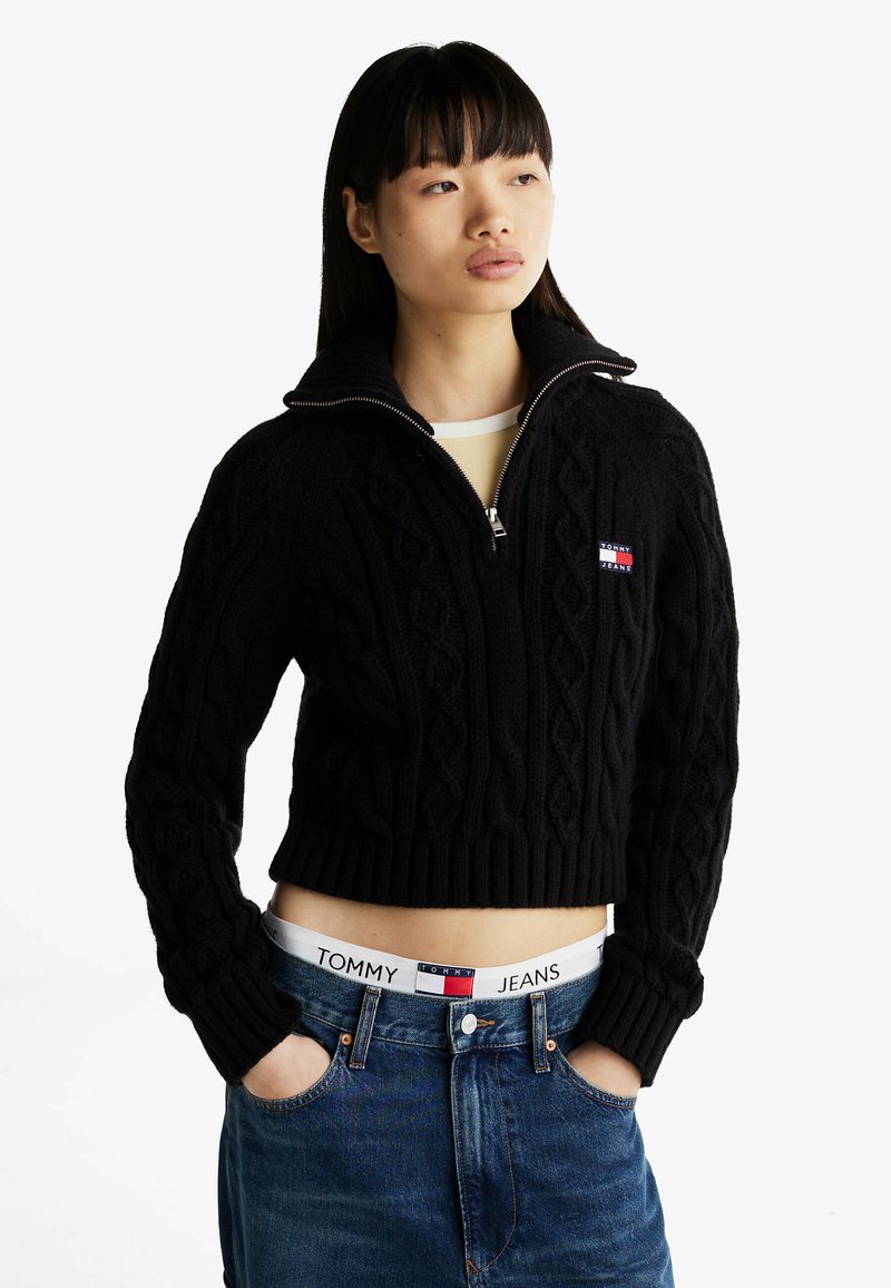 Maglione corto nero a trecce con colletto a mezzo zip, caratterizzato da una piccola toppa con logo. Abbinato a jeans a vita alta con marchio visibile sul cinturino.