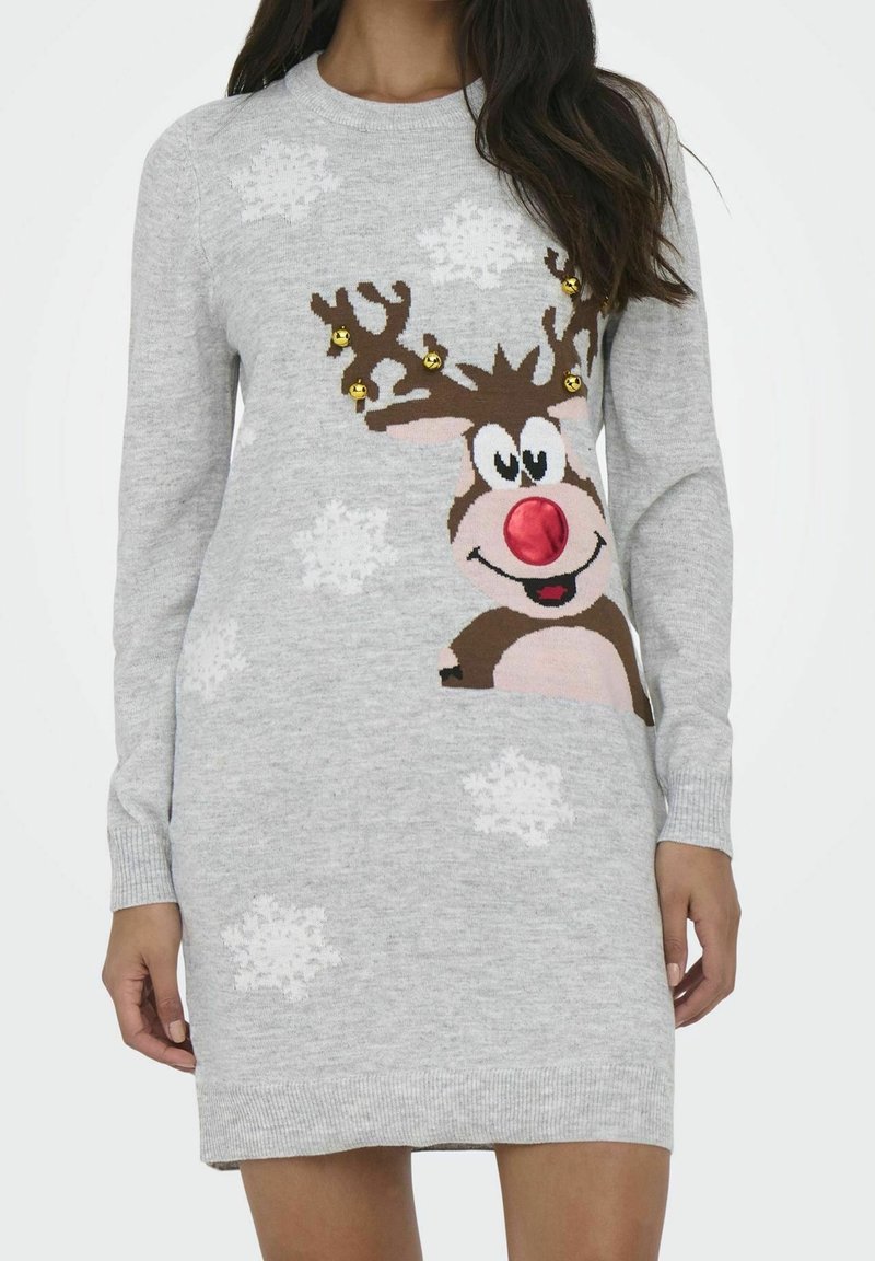 Femme portant une robe-pull grise avec un motif de renne souriant, un nez rouge, des grelots dorés sur les bois et des flocons de neige blancs.