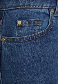 Blå denimjeans med en liten framficka, guldfärgade nitar, kontrasterande sömmar och en mjuk, texturerad ytfinish.