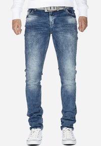 Jean en denim bleu avec un lavage délavé, coupe slim et détails légèrement usés. Associé à une ceinture blanche et des baskets blanches.