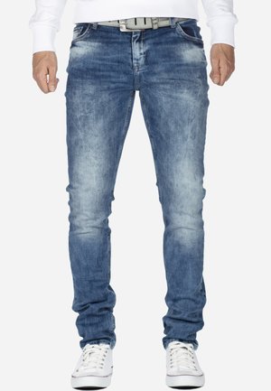 Jean en denim bleu avec un lavage délavé, coupe slim et détails légèrement usés. Associé à une ceinture blanche et des baskets blanches.
