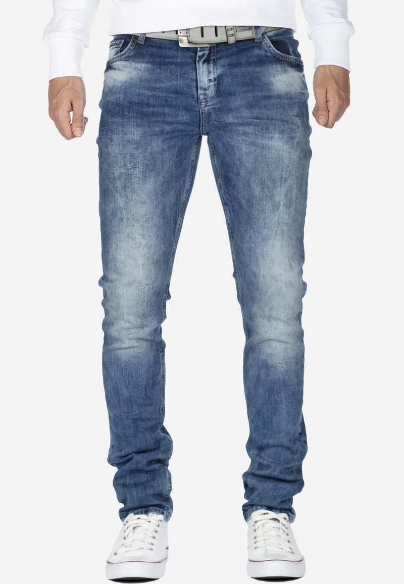 Jean en denim bleu avec un lavage délavé, coupe slim et détails légèrement usés. Associé à une ceinture blanche et des baskets blanches.