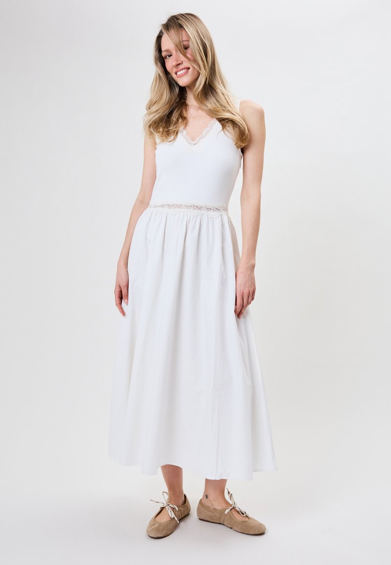 Femme aux longs cheveux blonds portant une robe blanche sans manches avec bordure en dentelle et des ballerines beiges à lacets, debout devant un fond uni.