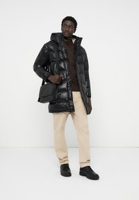 Black puffer coat with a hood, glossy finish, paired with a brown sweater, beige pants, and black shoes. Model carries a black shoulder bag.
