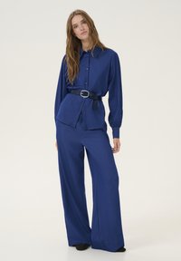 Camicia blu a maniche lunghe con bottoni, abbinata a pantaloni a gamba larga dello stesso materiale e colore. Include una cintura nera con fibbia argento.