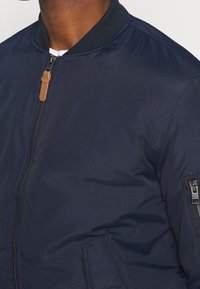 Giacca bomber della marina in tessuto liscio con colletto a coste, chiusura con zip e tasche laterali; presenta una tasca con zip sul manico sinistro.