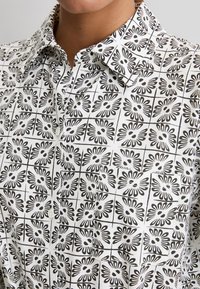 Camisa blanca de botones con un patrón de cuadrícula floral negro y un bolsillo en el pecho, usada por una persona cuyo cuello y barbilla son visibles.