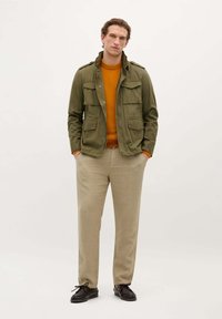 Olijfgroene militaire stijljas met twee borstzakken, oranje trui en beige broek, compleet met zwarte veterschoenen.