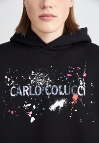 Čierna mikina s kapucňou a šnúrkou, s farebným dizajnom rozliatej farby a textom "CARLO COLUCCI" v tučnom písme.