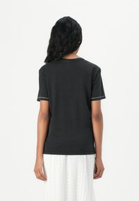 T-shirt en coton noir à manches courtes, avec des coutures contrastées aux poignets. La vue de dos montre un design simple et sans embellissement.