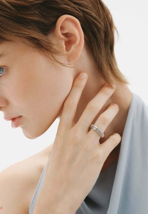 Mujer con cabello corto castaño tocándose el cuello, lleva un anillo plateado texturizado en el dedo medio y una blusa azul claro.