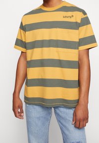 Žluté a olivově zelené pruhované tričko s kulatým výstřihem, krátkými rukávy a přední kapsou. Obsahuje logo Levis na hrudi.