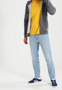 Sudadera gris con cremallera y forro interior blanco, camiseta de cuello redondo amarilla, vaqueros azul claro y zapatillas marinas con rayas blancas.