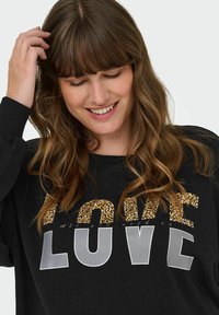 Sieviete smaida, skatās lejup, pieskaroties saviem matiem, ģērbusies melnā džempā ar uzrakstu "LOVE" pelēkās un leopardu raksta burtiem.