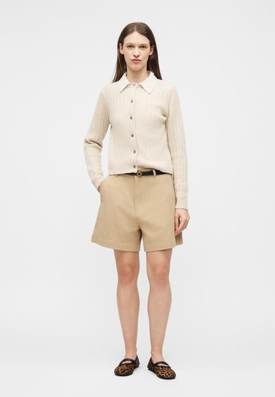 Femme debout portant un cardigan beige côtelé boutonné avec un col blanc, un short beige mi-genoux, une ceinture noire et des ballerines à imprimé léopard.