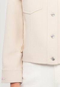 Giacca corta beige chiaro realizzata in tessuto morbido. Presenta due tasche sul petto, bottoni argentati e un sottile etichetta rosa sul polsino. Texture liscia.