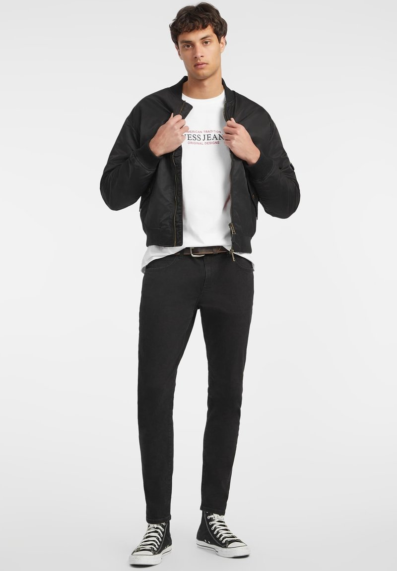 Veste aviateur noire avec fermeture éclair, portée sur un t-shirt blanc, associée à un jean noir et des baskets montantes noires.