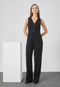 Crni krojeni kombinezon s kratkim, bez rukava, gornjim dijelom koji se zakopčava na dugmad, uz struk koji pristaje i široke nogavice. Glatka tkanina.