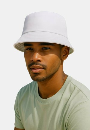 Homme à la barbe courte portant une chemise vert clair et un chapeau bob blanc, regardant légèrement hors caméra avec une expression neutre sur un fond uni.