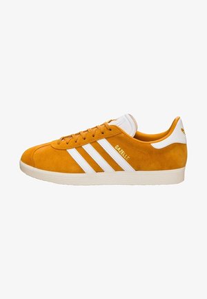 Pomarańczowy zamszowy sneaker Adidas Gazelle z białymi paskami, białym elementem na pięcie i beżową gumową podeszwą widziany z boku.