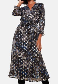 Černé květinové midi šaty s dlouhými volánovými rukávy, výstřihem do V a páskem v pase, které nosí stojící osoba s jednou rukou zvednutou k vlasům.