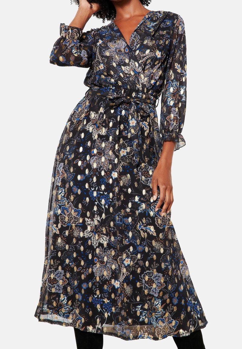Černé květinové midi šaty s dlouhými volánovými rukávy, výstřihem do V a páskem v pase, které nosí stojící osoba s jednou rukou zvednutou k vlasům.