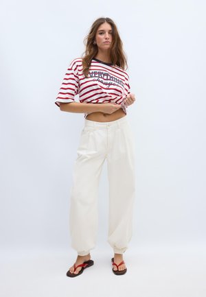 Frau mit langen braunen Haaren, die ein rot-weiß gestreiftes Crop-Top, cremefarbene weite Hosen und rote Flip-Flops trägt, steht vor einem schlichten weißen Hintergrund.