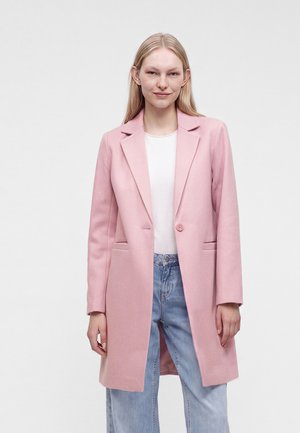 Femme aux longs cheveux blonds portant un manteau rose clair à un seul bouton, un haut blanc et un jean bleu clair, debout devant un fond blanc uni.