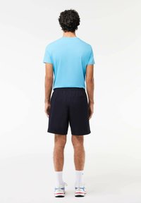 Camicia azzurra chiara a maniche corte, abbinata a pantaloni corti scuri, indossata con calzini bianchi e sneakers, vista da dietro. Design semplice e sportivo.