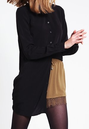 Femme portant une chemise longue noire boutonnée sur un short beige orné de dentelle et un collant foncé, les mains jointes à la taille.