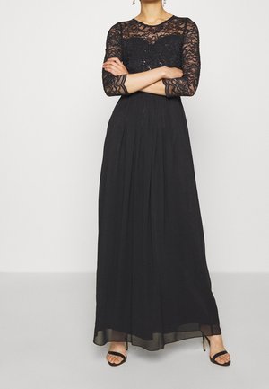 Femme portant une robe noire longue au sol avec des manches trois-quarts en dentelle et des détails en paillettes, associée à des talons noirs ouverts à l'avant.