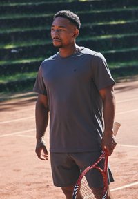 Mann i grå treningsklær som holder en rød tenniskølle, står på en leirtennisbane med grønne tribuner i bakgrunnen.