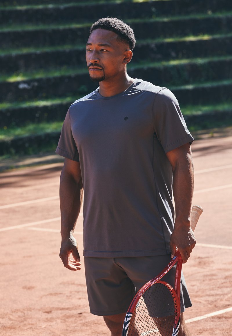 Mann i grå treningsklær som holder en rød tenniskølle, står på en leirtennisbane med grønne tribuner i bakgrunnen.