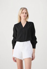 Schwarze Langarmbluse mit V-Ausschnitt und dezenten Falten, kombiniert mit weißen Jeans-Shorts mit ausgefranstem Saum. Glatter Stoff und lockere Passform.