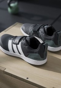 Black athletic shoes with white stripes and green accents, featuring a textured upper, flat sole, and adjustable strap across the instep.