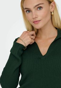 Grön ribbad tröja med V-ringning. Modellen bär guldsmycken, inklusive ett armband, som visar glatt textur och en figurnära design.