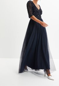 Mörkblå aftonklänning med trekvarts ärmar i spets, utsmyckad bodice och en flödande, genomskinlig tylskjol. Höga vita skor synliga.