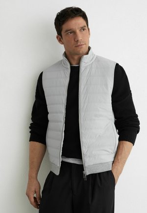 Uomo che indossa un gilet trapuntato grigio chiaro sopra una maglia nera a maniche lunghe, in piedi contro un muro bianco, guarda di lato.