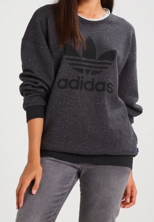 Kobieta w ciemnoszarej, nakrapianej bluzie Adidas z dużym czarnym logo, w połączeniu z szarymi dżinsami, stojąca z jedną ręką swobodnie opuszczoną wzdłuż ciała.