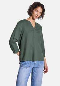 Blusa in lino verde con scollo a V, maniche a tre quarti e vestibilità rilassata, abbinata a jeans in denim blu chiaro.