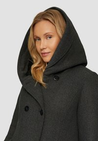 Grijze wollen jas met een grote capuchon, dubbele knoopsluiting en een gestructureerde stof. Kenmerken zijn onder andere twee zwarte knopen en een getailleerde pasvorm.