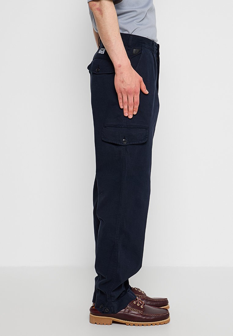 Granatowe spodnie cargo wykonane z bawełny, z bocznymi kieszeniami z zapięciem na guziki i luźnym krojem, zestawione z ciemnobrązowymi butami.