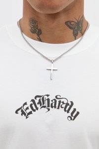 Biely tričko so výraznou čiernou grafikou "Ed Hardy". Strieborný náhrdelník s krížom a viditeľné květinové a motýlie tetovania.