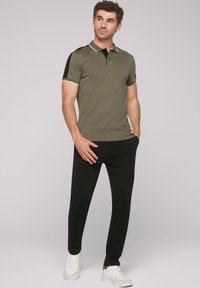 Olivgrünes Poloshirt mit schwarzen Ärmeln, weißer Paspelierung und einem Zwei-Knopf-Verschluss, kombiniert mit schwarzen Jogginghosen und weißen Sneakers.