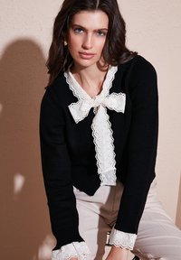 Black sweater featuring a white lace bow and collar, with lace accents on cuffs, ribbed fabric texture, and a fitted design.